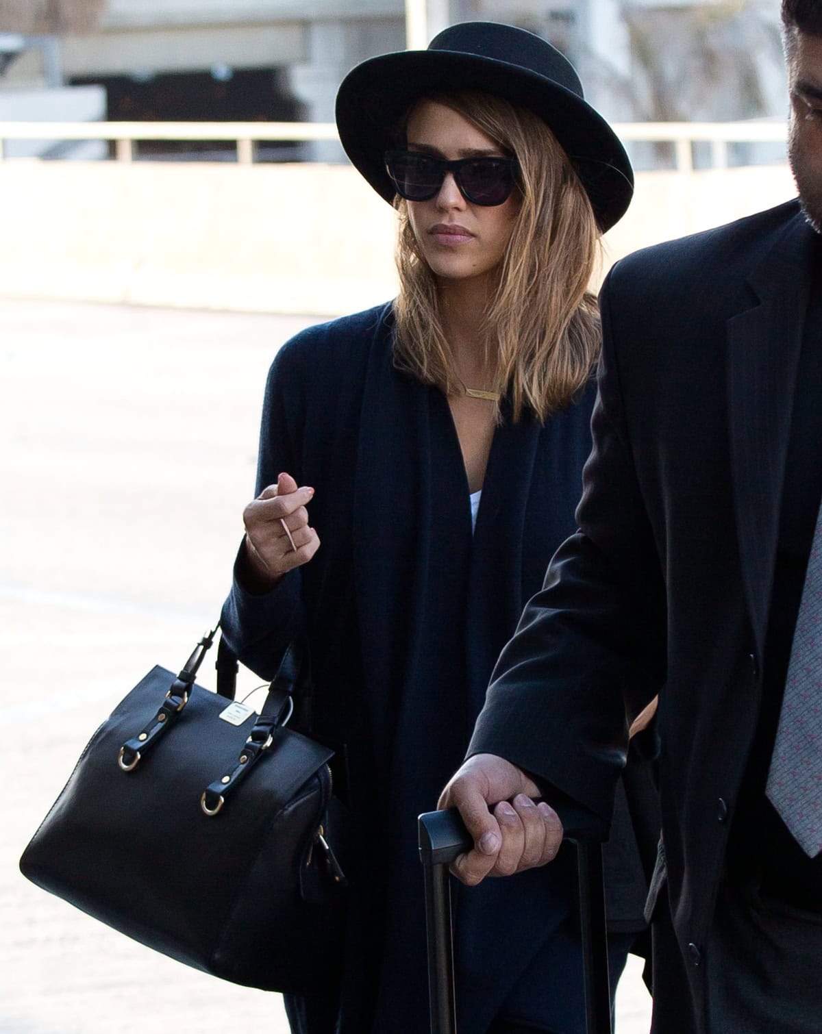 Jessica Alba arriving at LAX wearing a black wide-brim hat, dark sunglasses, and her hair styled in loose waves, carrying a sleek black handbag