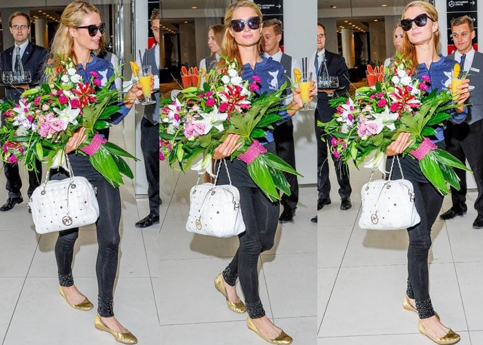 Paris Hilton juggles flowers, a fruity drink and a white tote bag as she arrives in Poland