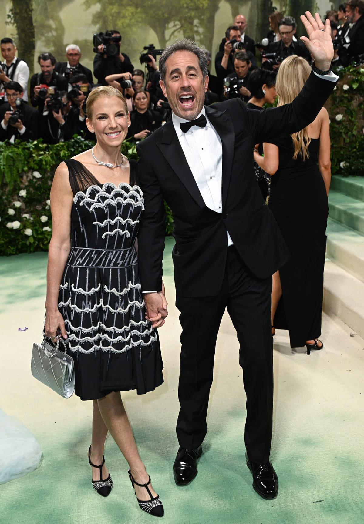 Jerry Seinfeld and his wife Jessica Seinfeld attend the Met Gala in style, with Jerry looking sharp in a classic black tuxedo while Jessica stuns in a chic black dress with intricate silver detailing, accessorized with a metallic handbag