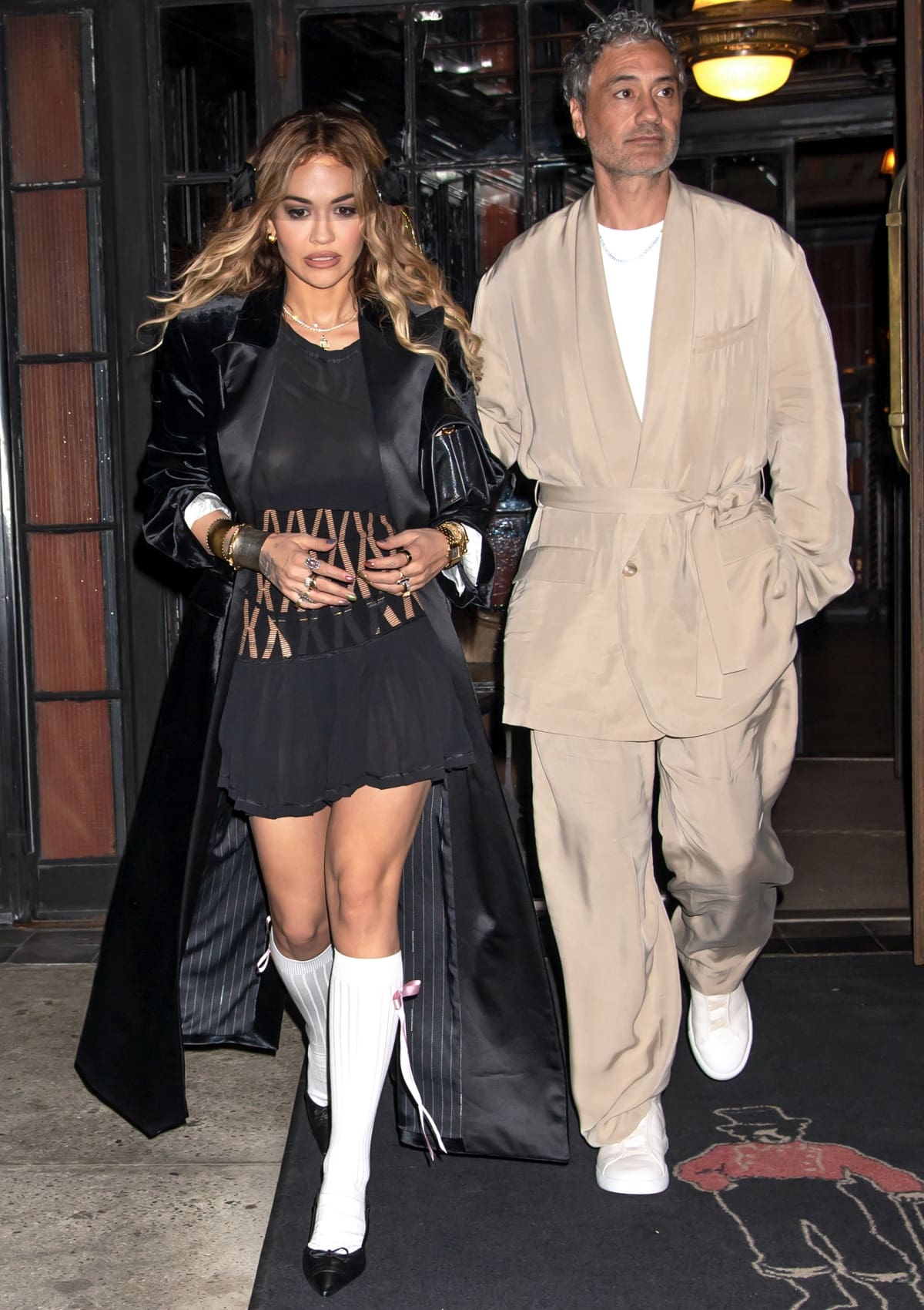 Taika Waititi wearing a beige coat, matching trousers, a crisp white T-shirt, and white trainers for a stylish contrast to wife Rita Ora’s daring look