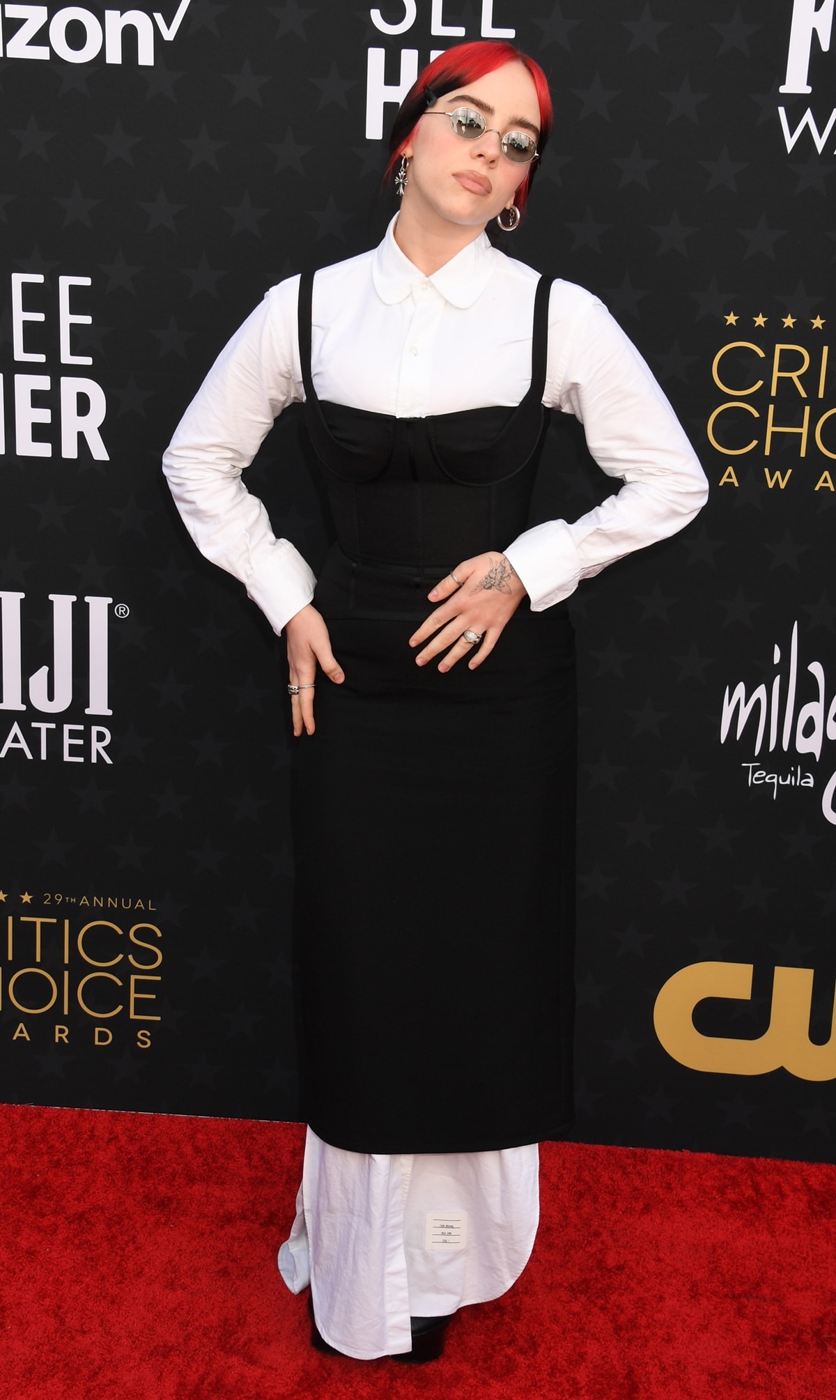 At the Critics Choice Awards at Barker Hangar on January 14, 2024 in Santa Monica, California, Billie Eilish donned a distinctive Thom Browne ensemble, consisting of a black formfitting corset-style dress over a floor-length white collared shirt, complemented by round glasses and her bright red and black hair tied back