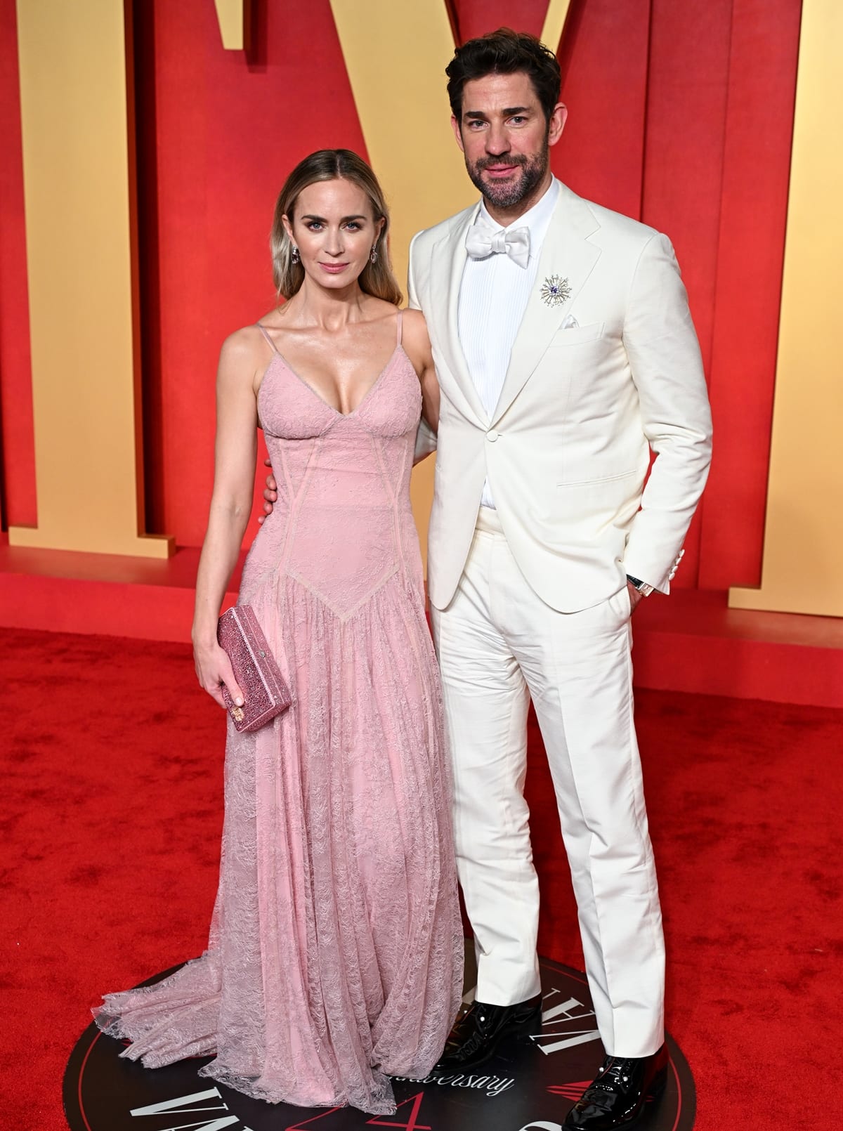 At the Vanity Fair Oscar Party, Emily Blunt switches to a custom-made pale pink lace gown by Dolce & Gabbana, playfully nodding to the Barbie movie, with John Krasinski complementing her look in a classic cream tuxedo, exemplifying Hollywood glamour and sophistication