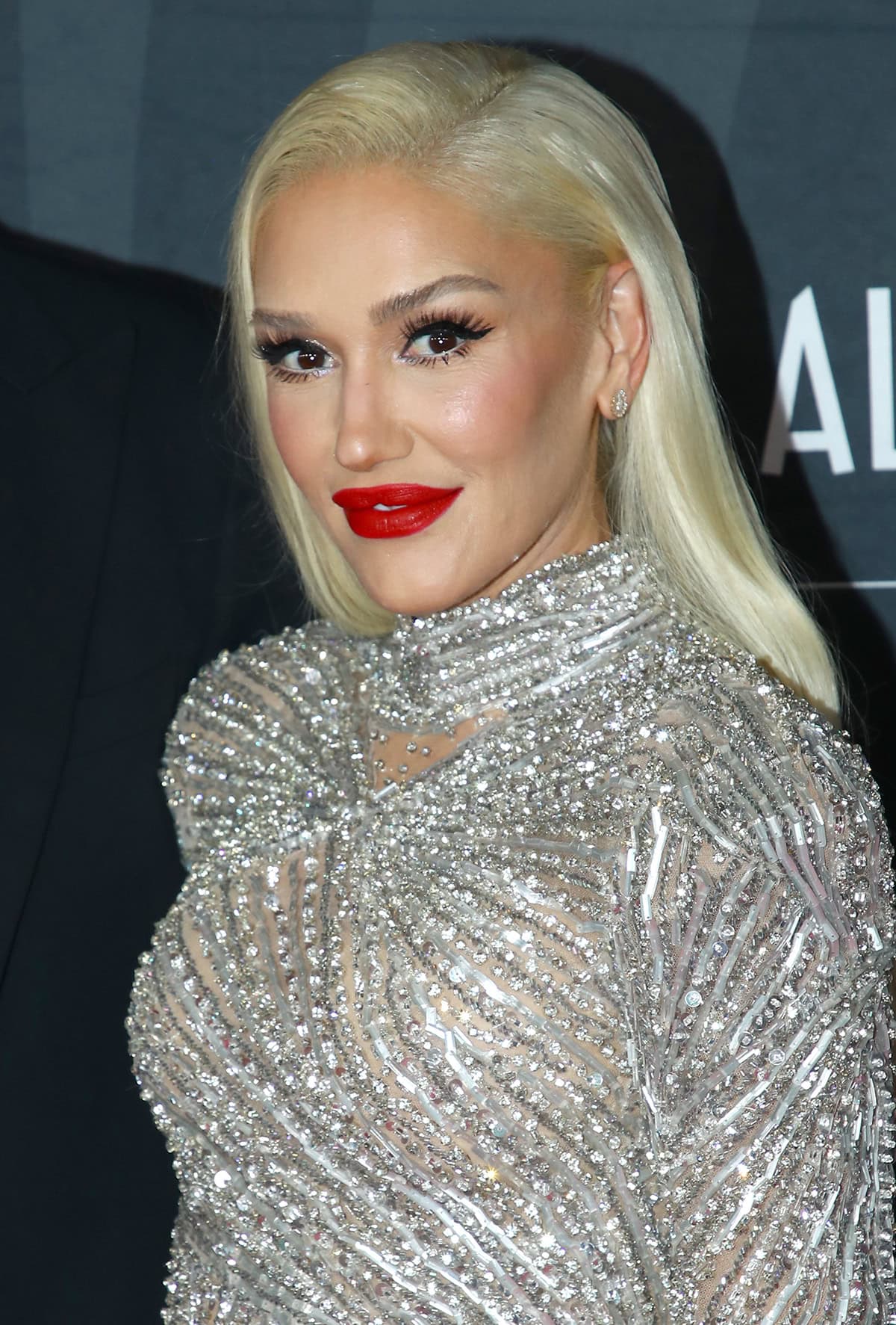 Gwen Stefani styles her platinum blonde hair straight with a side part and wears her signature red lipstick with bronze eyeshadow and fake eyelashes