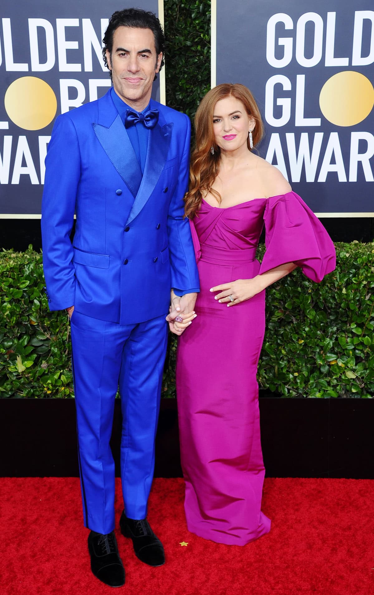 Isla Fisher stuns in a bold magenta gown with dramatic sleeves, while Sacha Baron Cohen complements her in a striking royal blue suit at the Golden Globe Awards