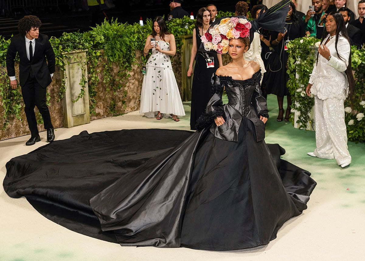 Zendaya's archival black Givenchy gown features a corset bodice, a lace-trimmed off-shoulder neckline, and an enormous skirt with a monarch train engulfing the Met Gala stairs