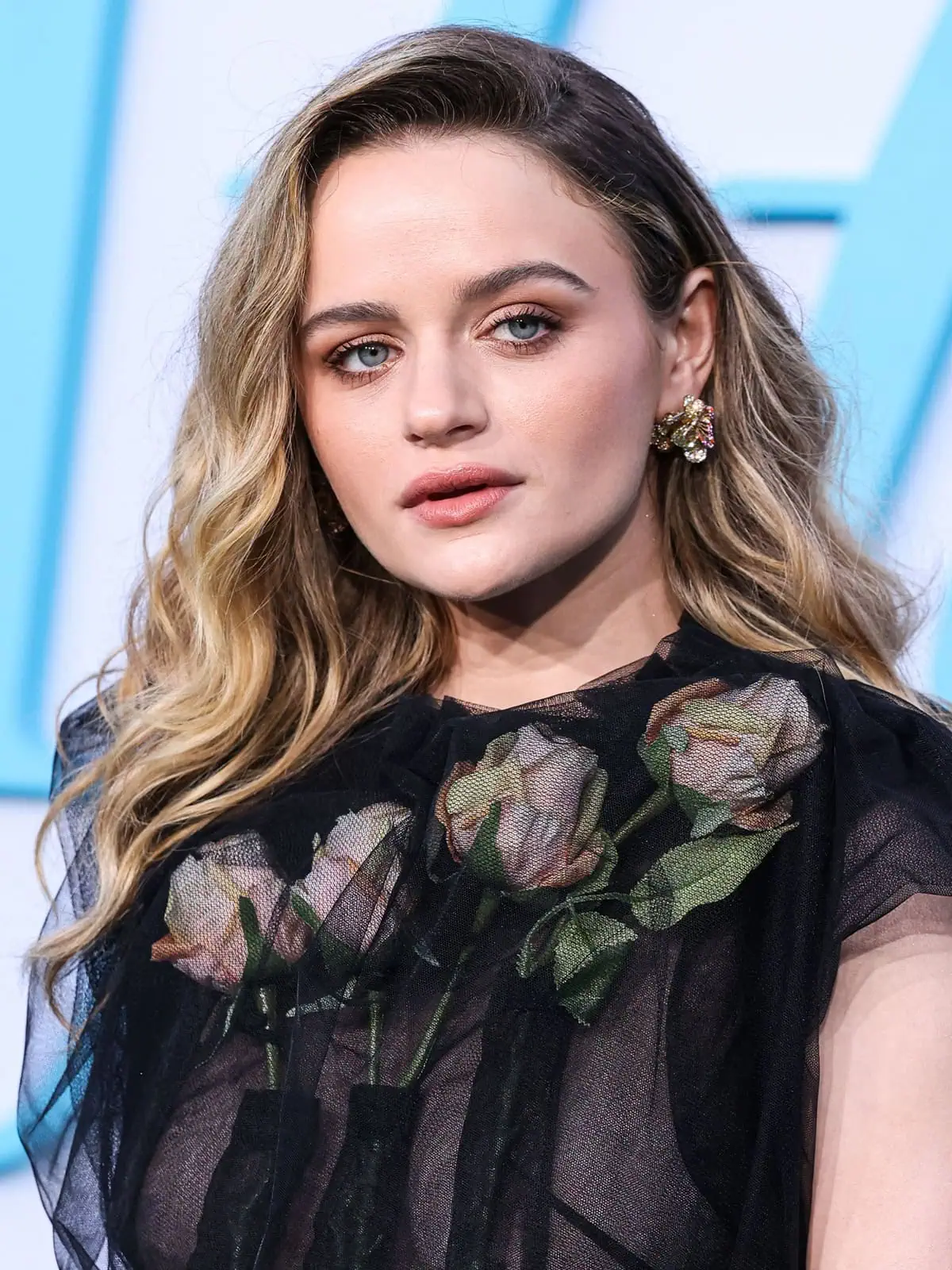 Joey King showcases her ethereal beauty with wavy blonde locks, minimal makeup, and delicate floral earrings, perfectly complementing her sheer black dress adorned with life-like roses