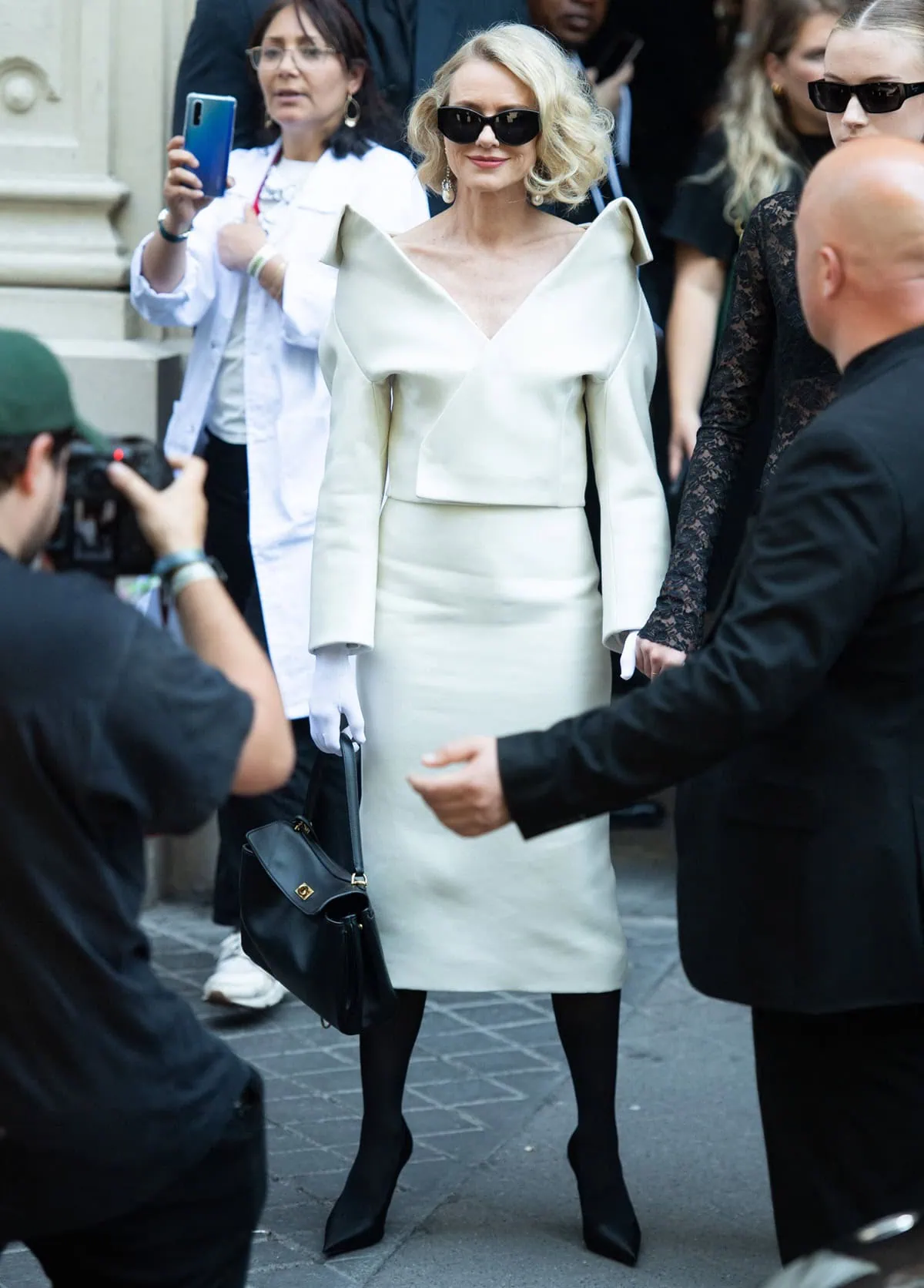 Naomi Watts dazzled at the Balenciaga show in a white skirt suit with a deep V neckline, paired with sheer black tights, black stilettos, matching white gloves, and statement diamond earrings