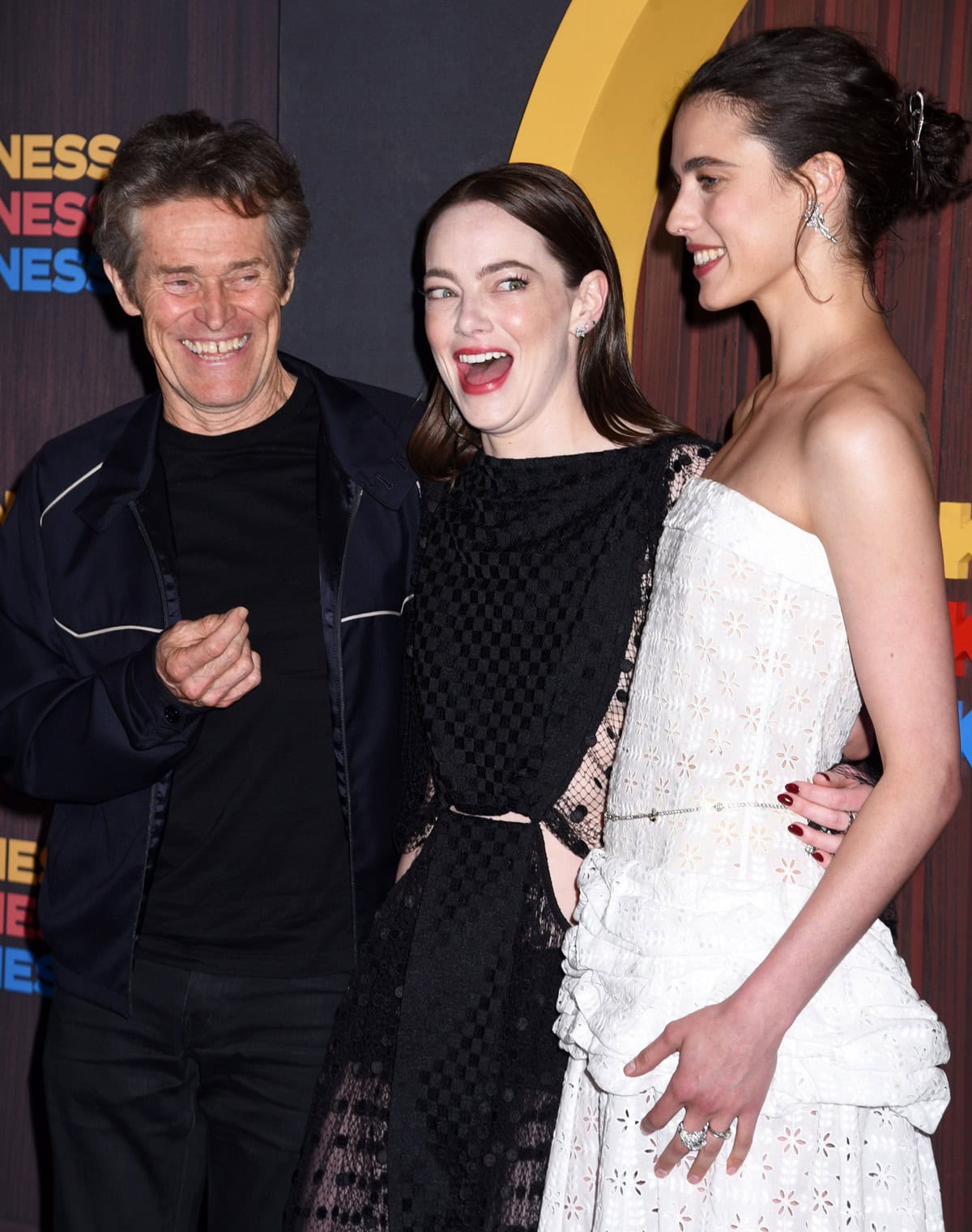 Emma Stone radiates joy with a wide smile and sparkling eyes, while Willem Dafoe beams with a hearty laugh, and Margaret Qualley smiles warmly, capturing a moment of genuine camaraderie and happiness among the trio
