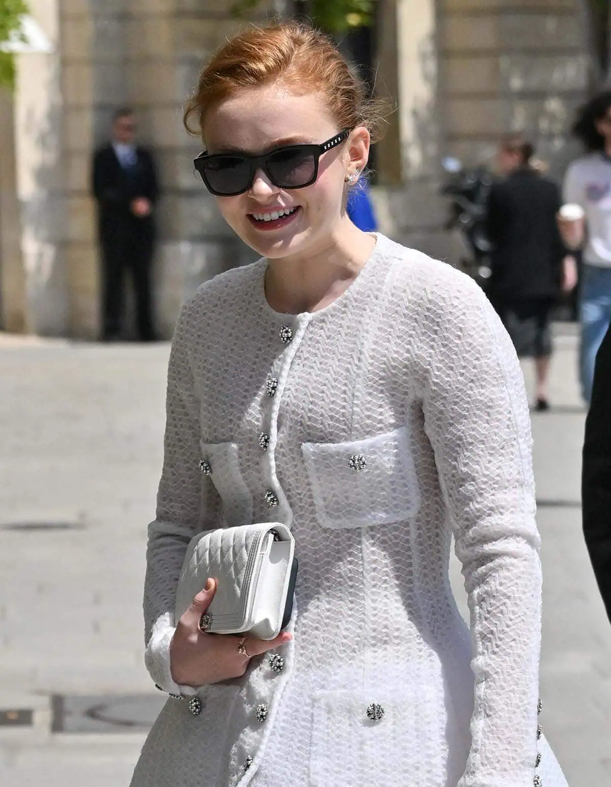 Sadie Sink accessorizes her white dress with white gold and yellow gold jewelry by Chanel, a white Chanel purse, and black sunnies