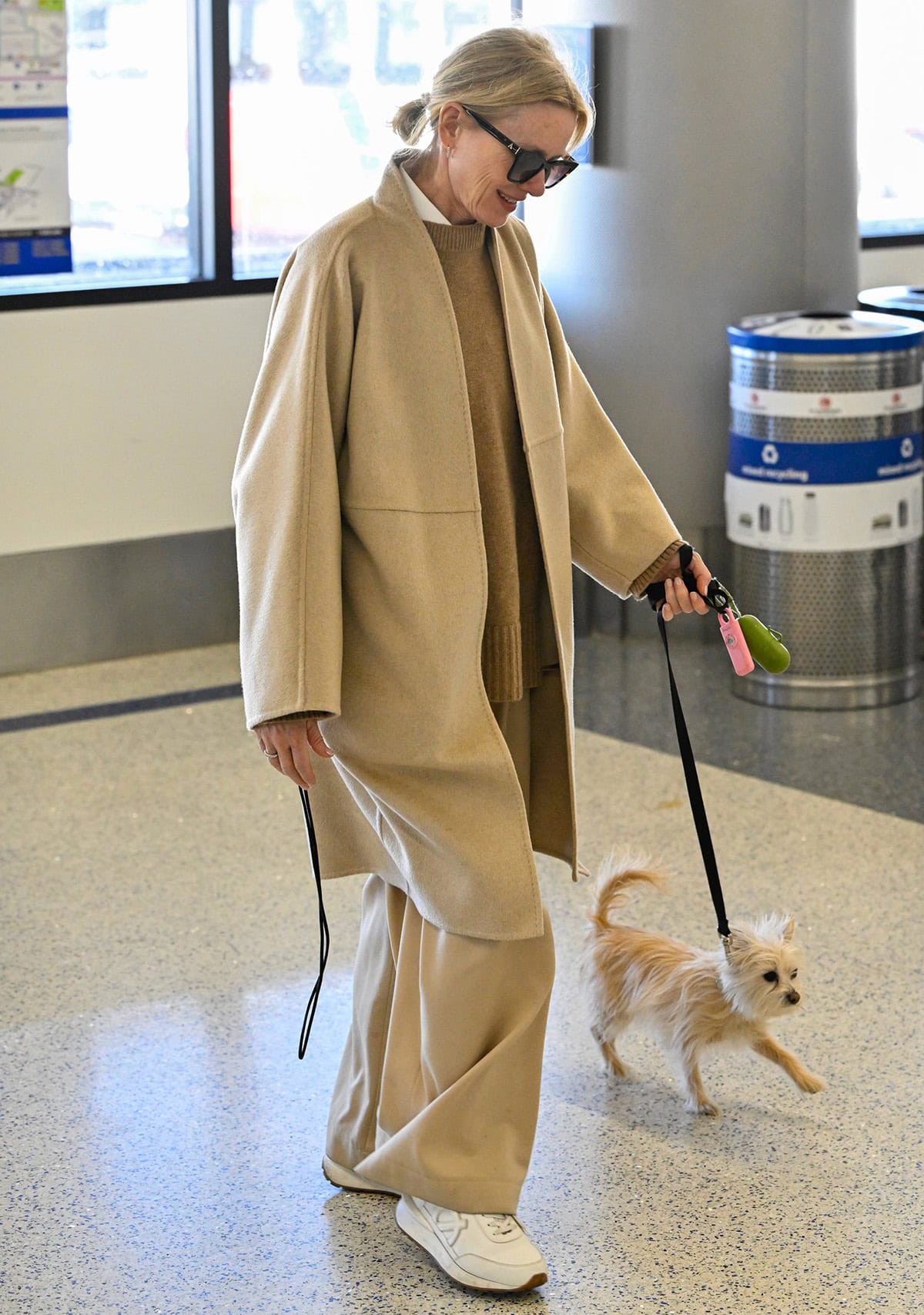 Naomi Watts' airport style includes cozy neutrals featuring a beige wool coat, matching baggy pants, a camel sweater, and a white shirt