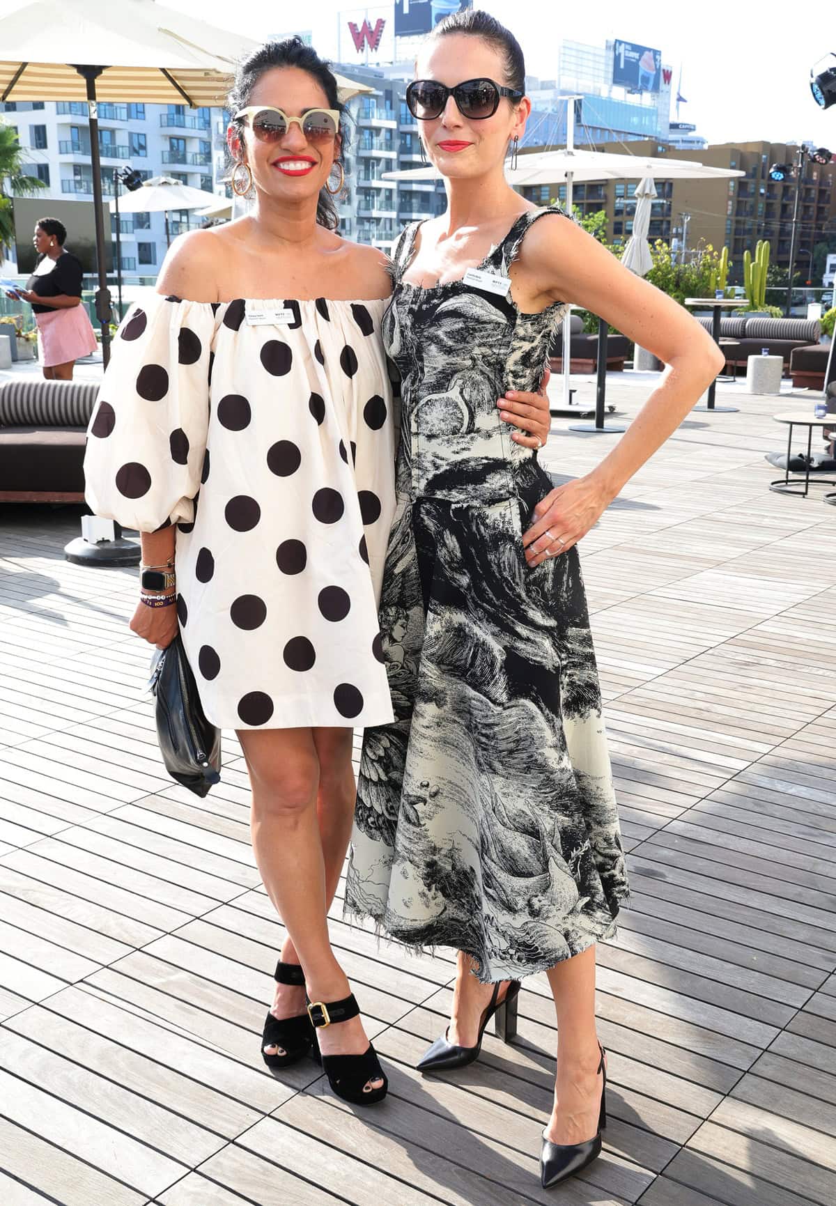 Tiffany Smith, wearing a playful polka-dot mini dress and platform sandals, stands beside the elegant Camilla Belle in a black-and-white printed midi dress and pointed heels, highlighting their stylish looks and slight height difference at the MPTF NexGen Annual Summer Party in Hollywood on June 23, 2024