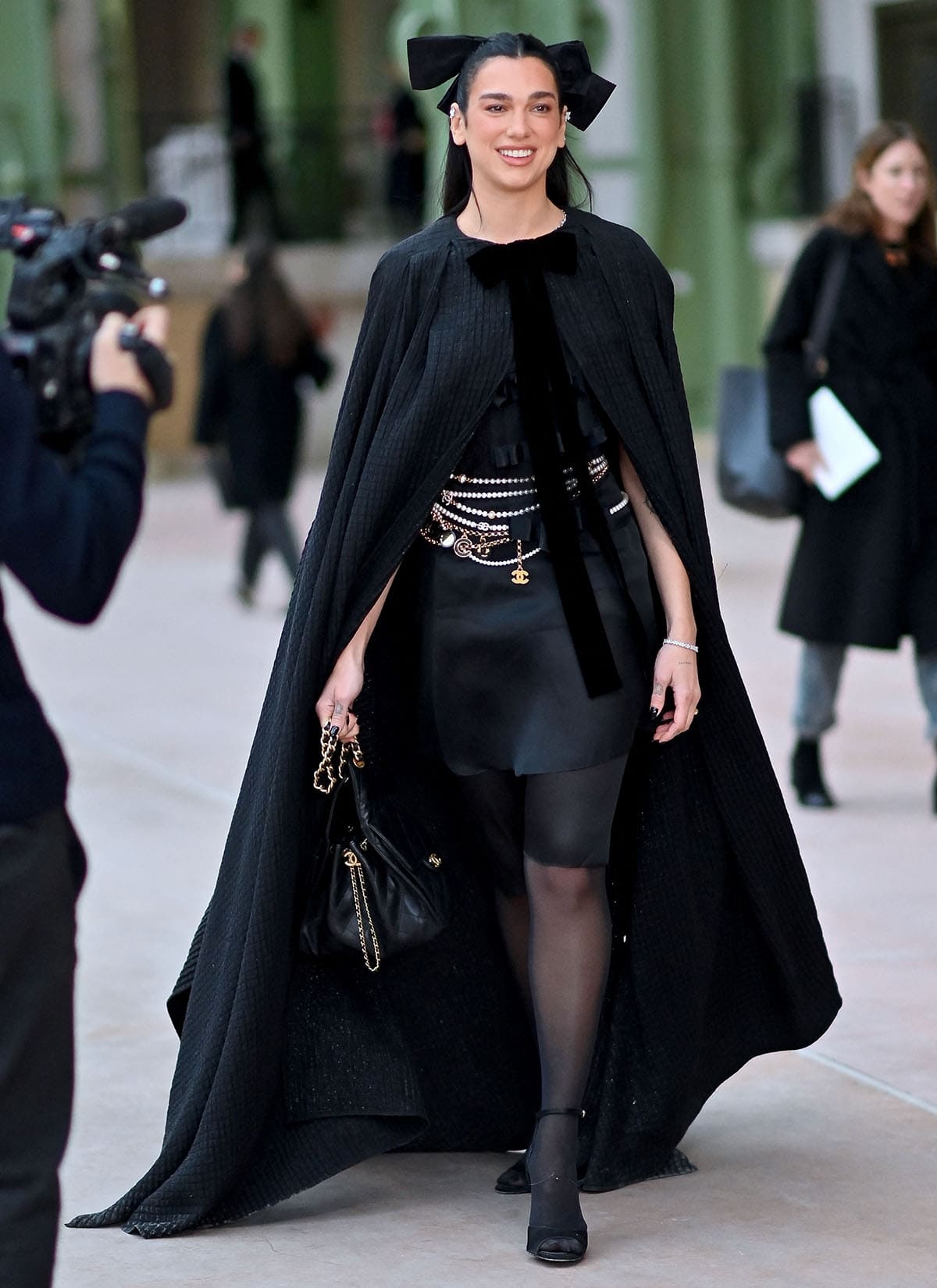 Dua Lipa exudes an enigmatic aura in her quilted black cape and black satin mini-dress with bow accents injecting a coquette charm to the look