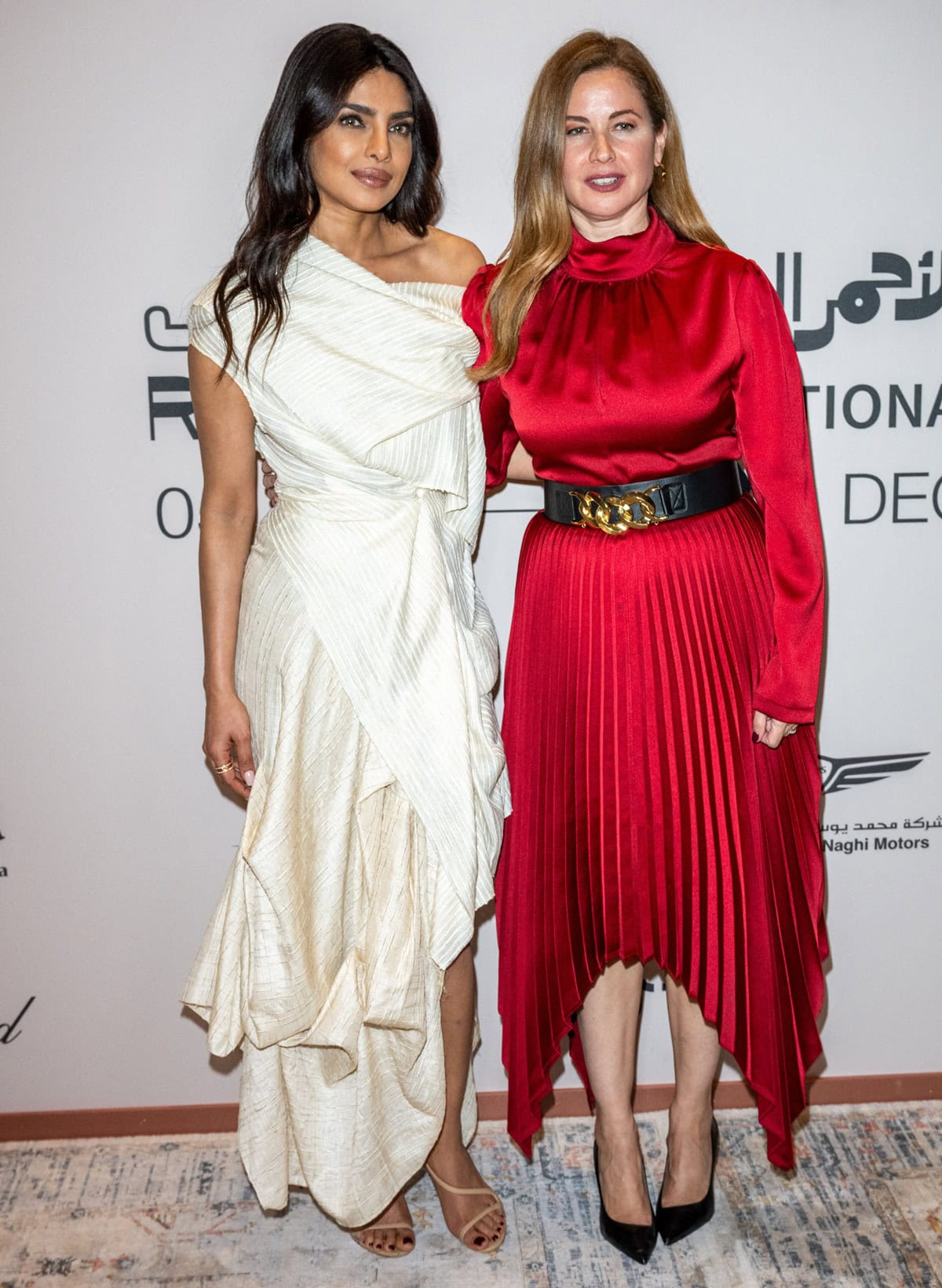 Priyanka Chopra Jonas stuns in an elegant off-white asymmetrical gown paired with strappy nude heels, while Raya Abirached captivates in a striking red pleated dress cinched with a statement belt, as they pose ahead of the In Conversation With Priyanka Chopra Jonas session at the Red Sea International Film Festival 2024 on December 11 in Jeddah, Saudi Arabia