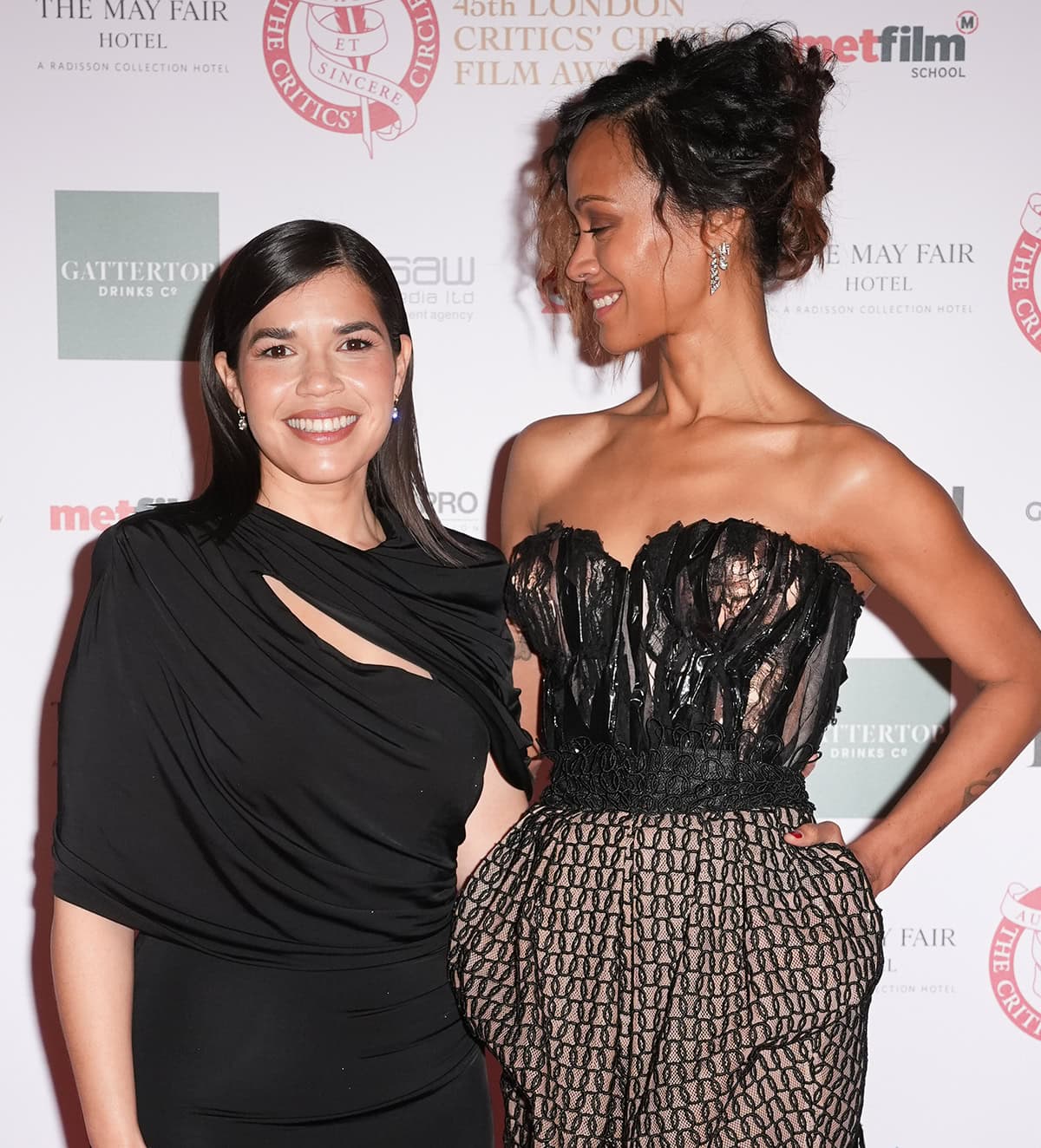 Zoe Saldana, towering over America Ferrera, exudes elegance in her avant-garde Maison Margiela dress, while America stuns in a sleek black gown; Zoe’s two-toned curly tresses styled in a messy updo complete her edgy look