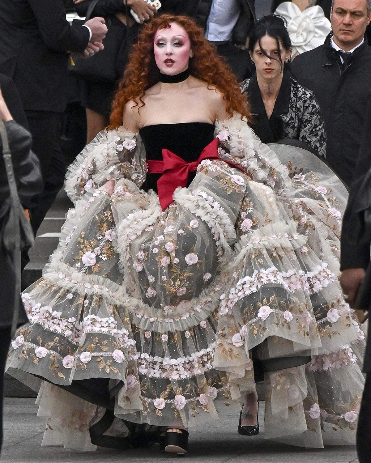 Chappell Roan steals the show in a Valentino tulle ball gown featuring a black corset bodice, a huge red ribbon, puffy tulle sleeves, and a voluminous skirt with 3D floral and leaf accents