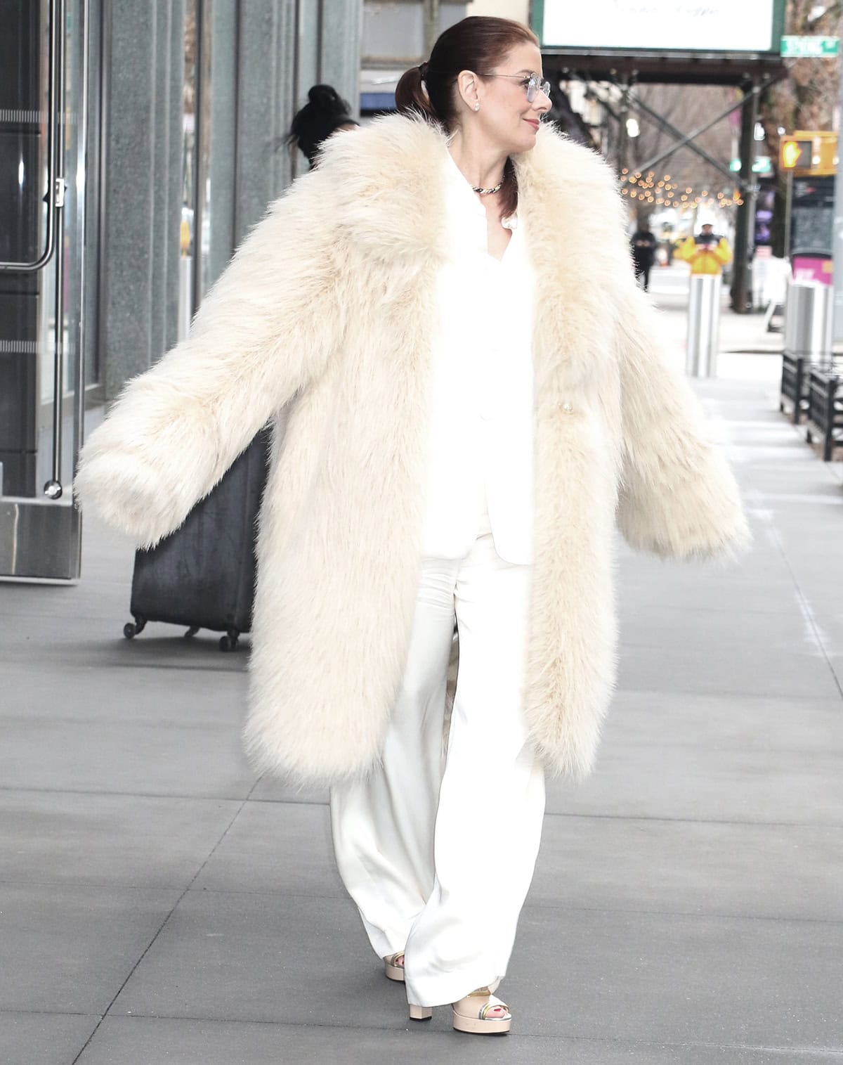 Debra Messing embraces her mafia boss-wife role by wearing an extravagant cream-colored fur coat over a white shirt and wide-leg pants combination