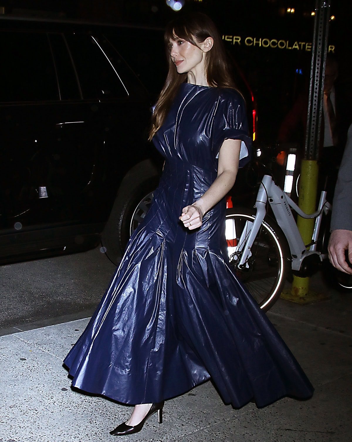 Elizabeth Olsen teams her black pumps with a navy PVC dress by Proenza Schouler featuring an empire drop waist, a bulky ruched bodice, and a long pleated skirt