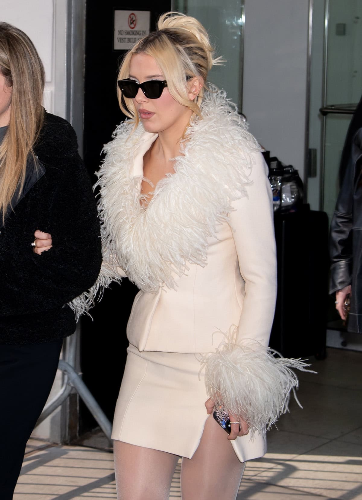 Millie Bobby Brown completes her elegant, feminine look with a cream-colored leather skirt suit with feather lapels, collar, and cuffs